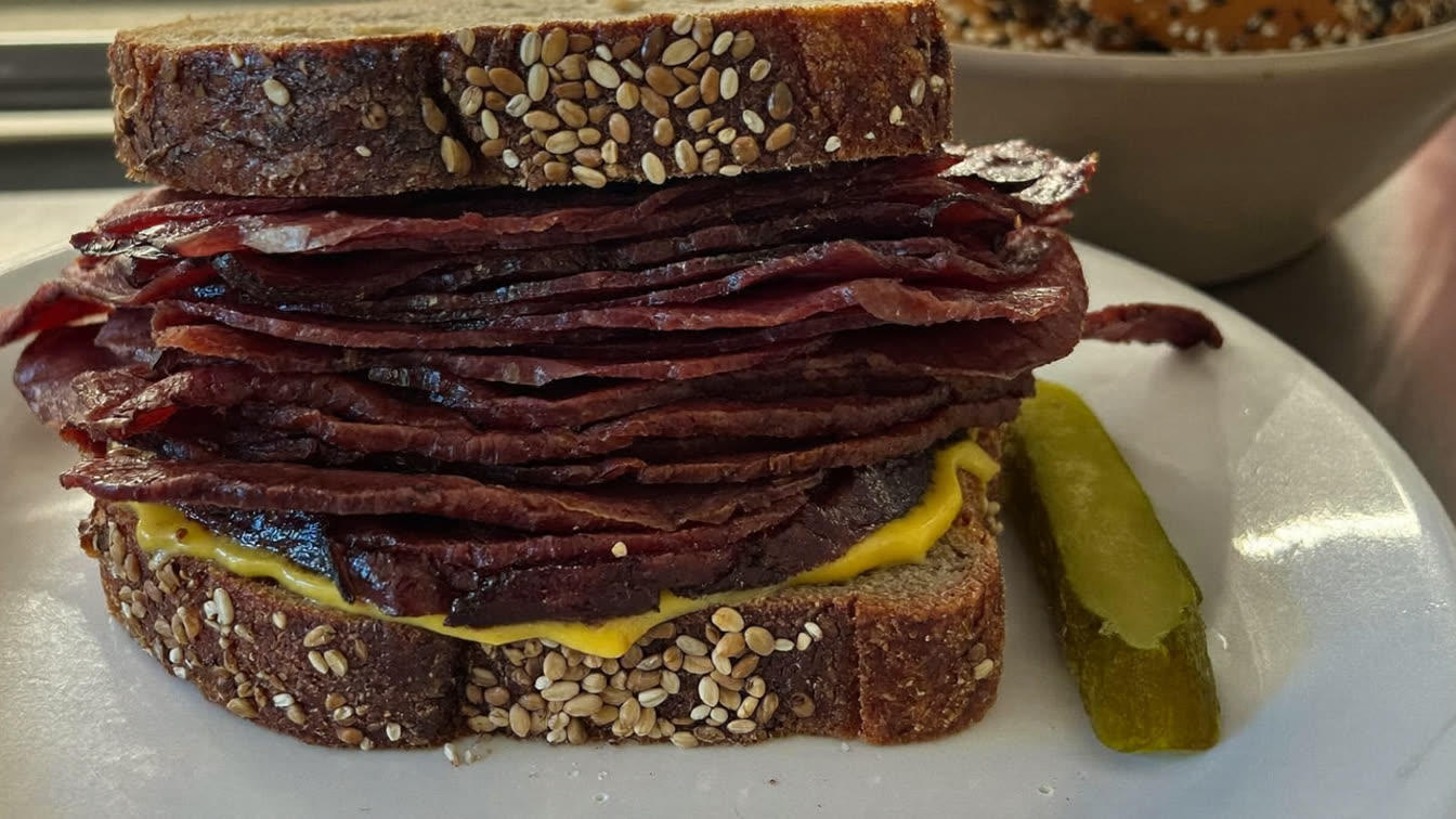 A towering Montreal-style smoked meat sandwich on seeded rye with mustard and a dill pickle spear