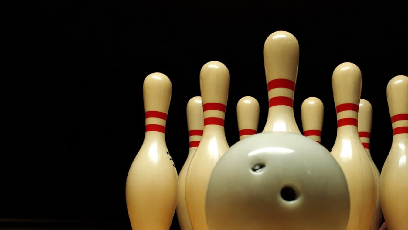 A bowling ball on a polished lane facing a rack of ten white bowling pins with red neck stripes