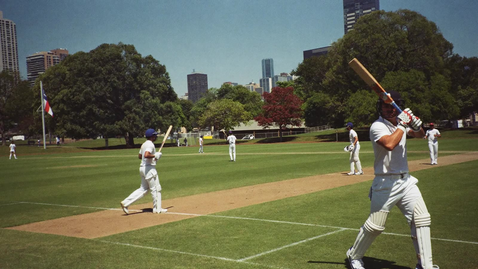 Batsman and fielders in white on a cricket pitch in a city park